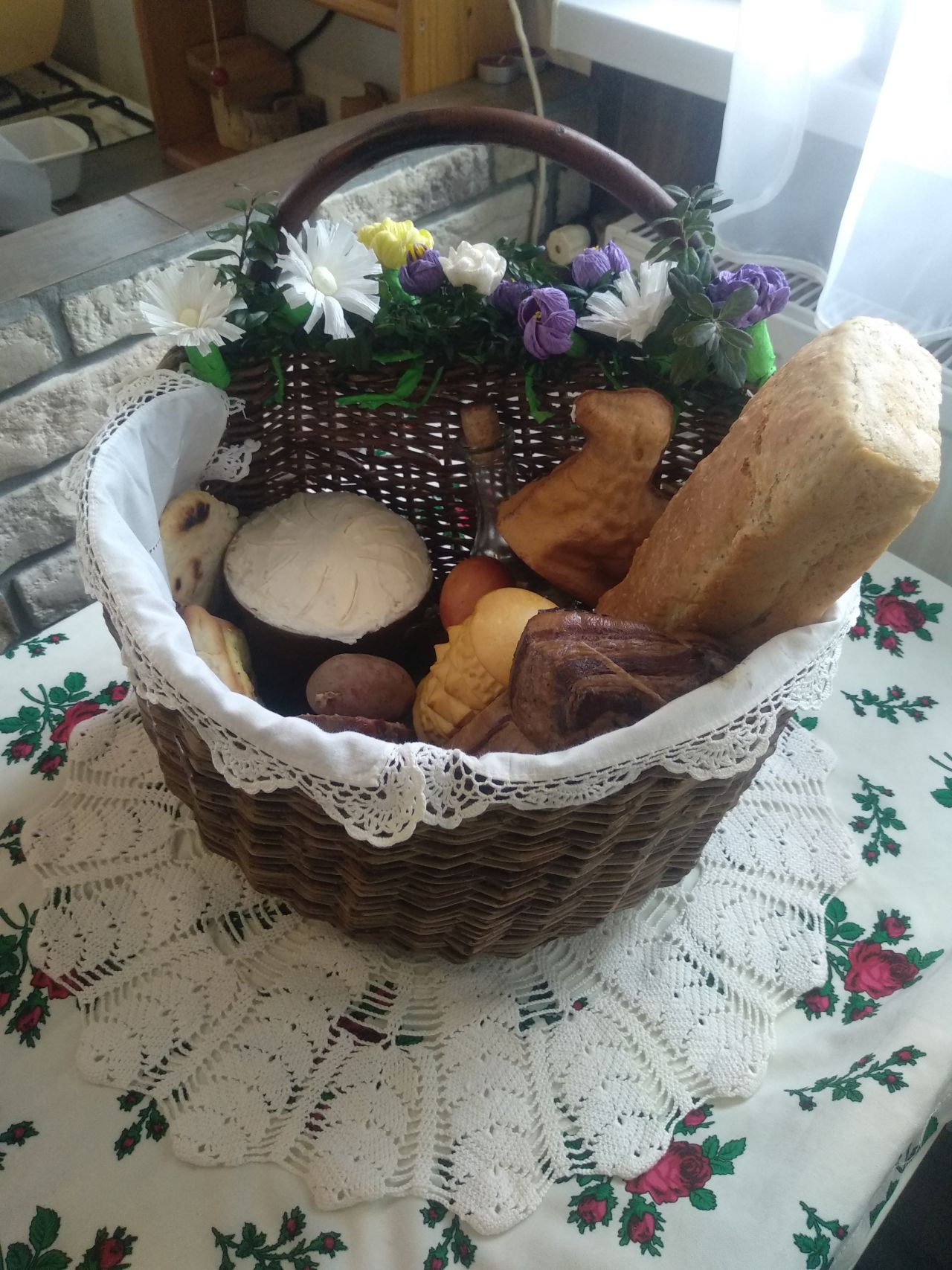 wiklinowy koszyk z białą serwetą, w środku chleb z formy, sery, jajko barwione na ciemnoczerwono, wędliny i inne wiktuały, szklana butelka z korkiem, całość przystrojona zielonymi gałązkami oraz białymi i fioletowymi kwiatami 