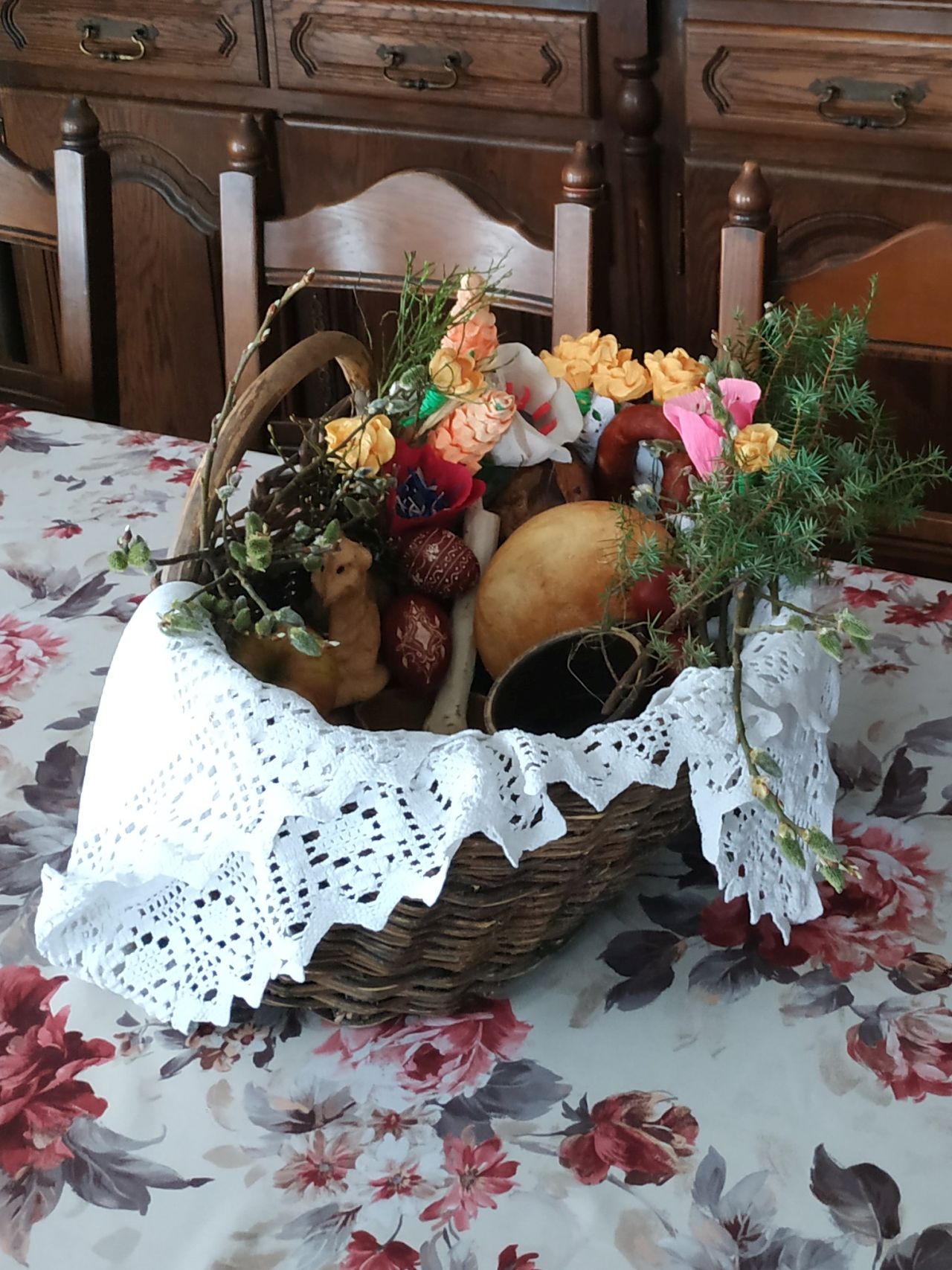 wiklinowy koszyk z białą, haklowaną serwetą, w środku baranek, chleb, pisanki ii inne wiktuały przystrojone w zielone gałązki oraz kolorowe kwiaty z bibuły