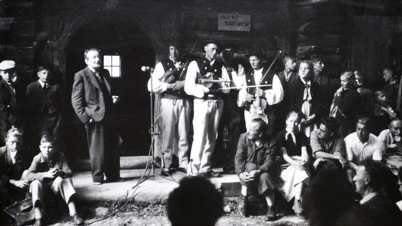 grupa ludzi stoi przed dworem, przemawia mężczyzna w garniturze, obok stoi zespół w strojach regionalnych