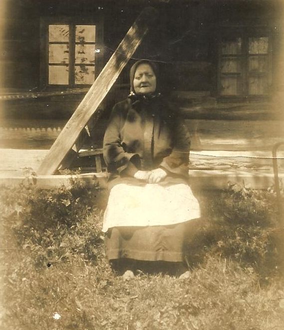stara, pożółkła, czarno-biała fotografia: starsza kobieta siedzi na krześle przed drewnianym budynkiem zwrócona w stronę obiektywu, na głowie ma zawiązaną chustkę, jest ubrana w ciemny strój z jasnym fartuchem