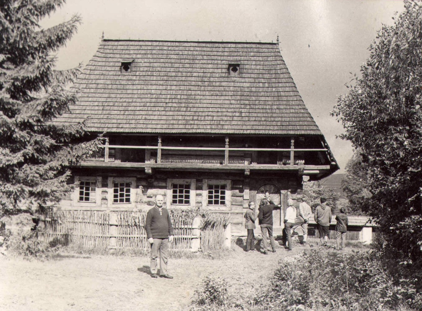 Andrzej Pilch stojący przed zabytkową drewnianą chałupą w skansenie, z nim, po prawej widoczni inni ludzie stojący przed drzwiami do chałupy