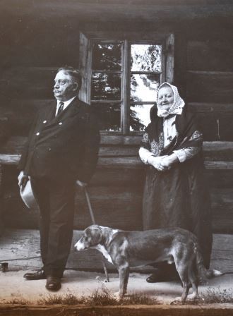 mężczyzna w garniturze z kapeluszem i laską, Sándor Lattyak, i kobieta w wiejskim stroju i chustce na głowie, Joanna Wilczkowa, stoją przed drewnianym dworem Moniaków